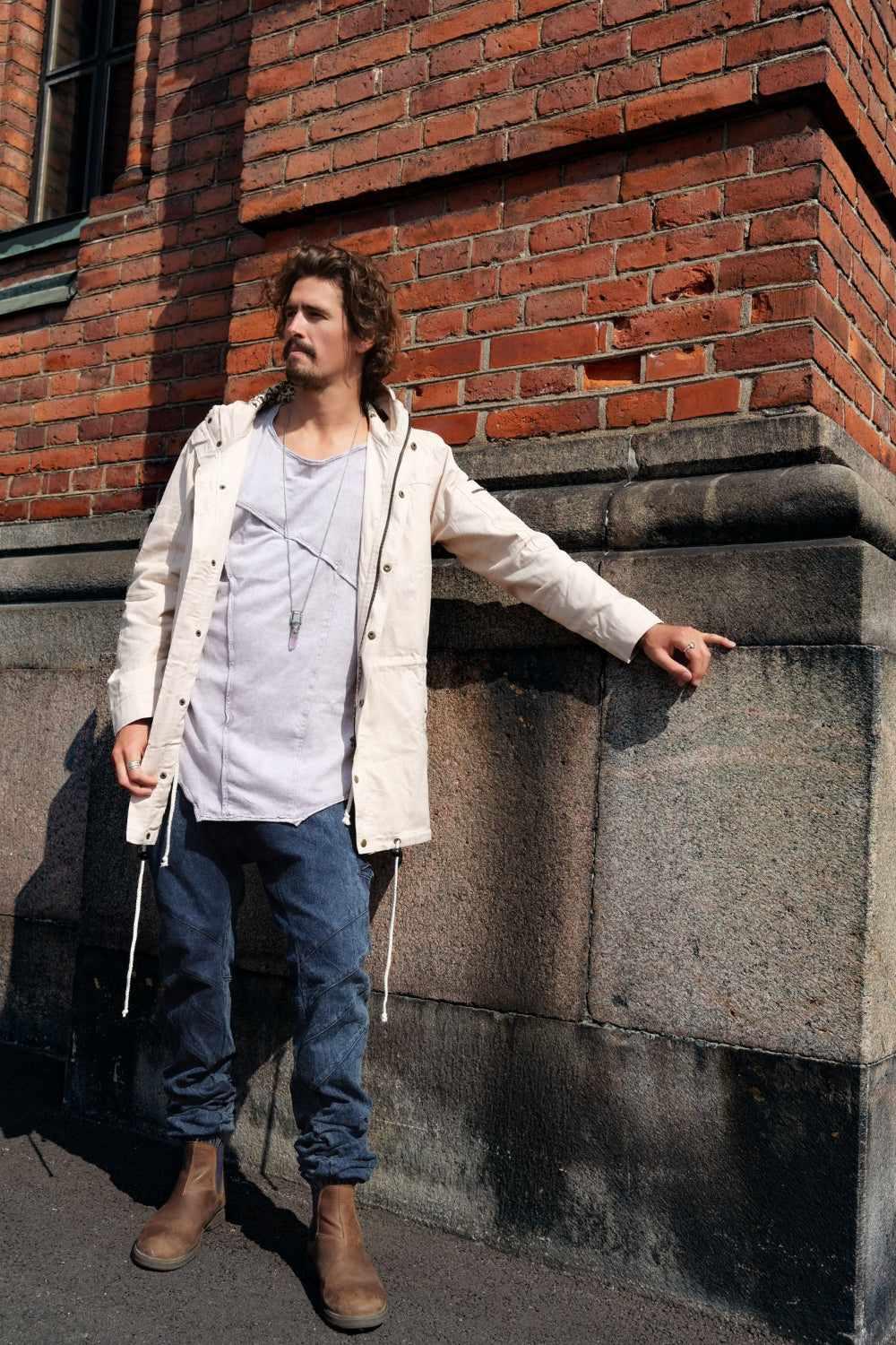Man leaning against a brick wall wearing a beige jacket and blue jeans.