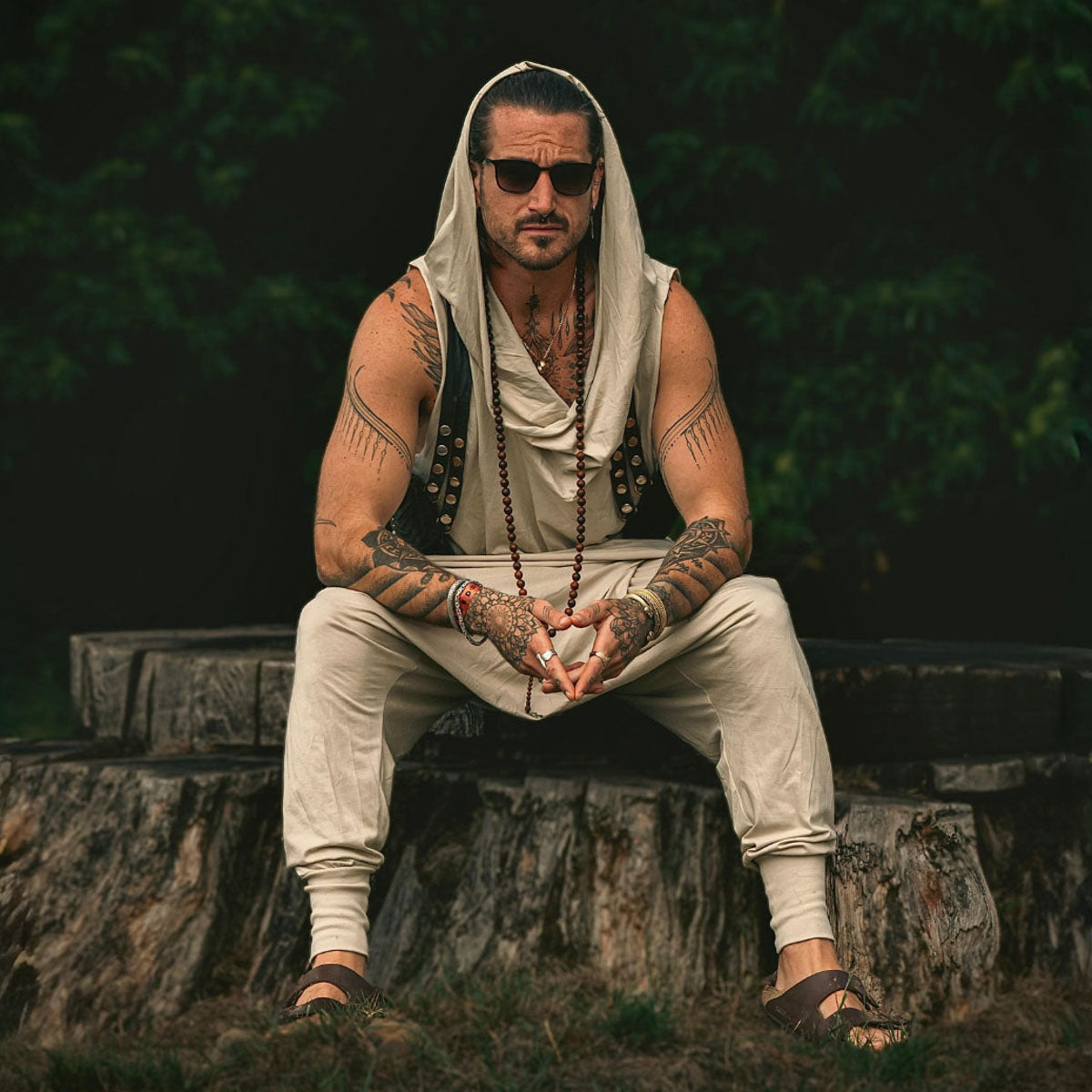 Man with tattoos and a white headscarf sitting on a tree stump outdoors