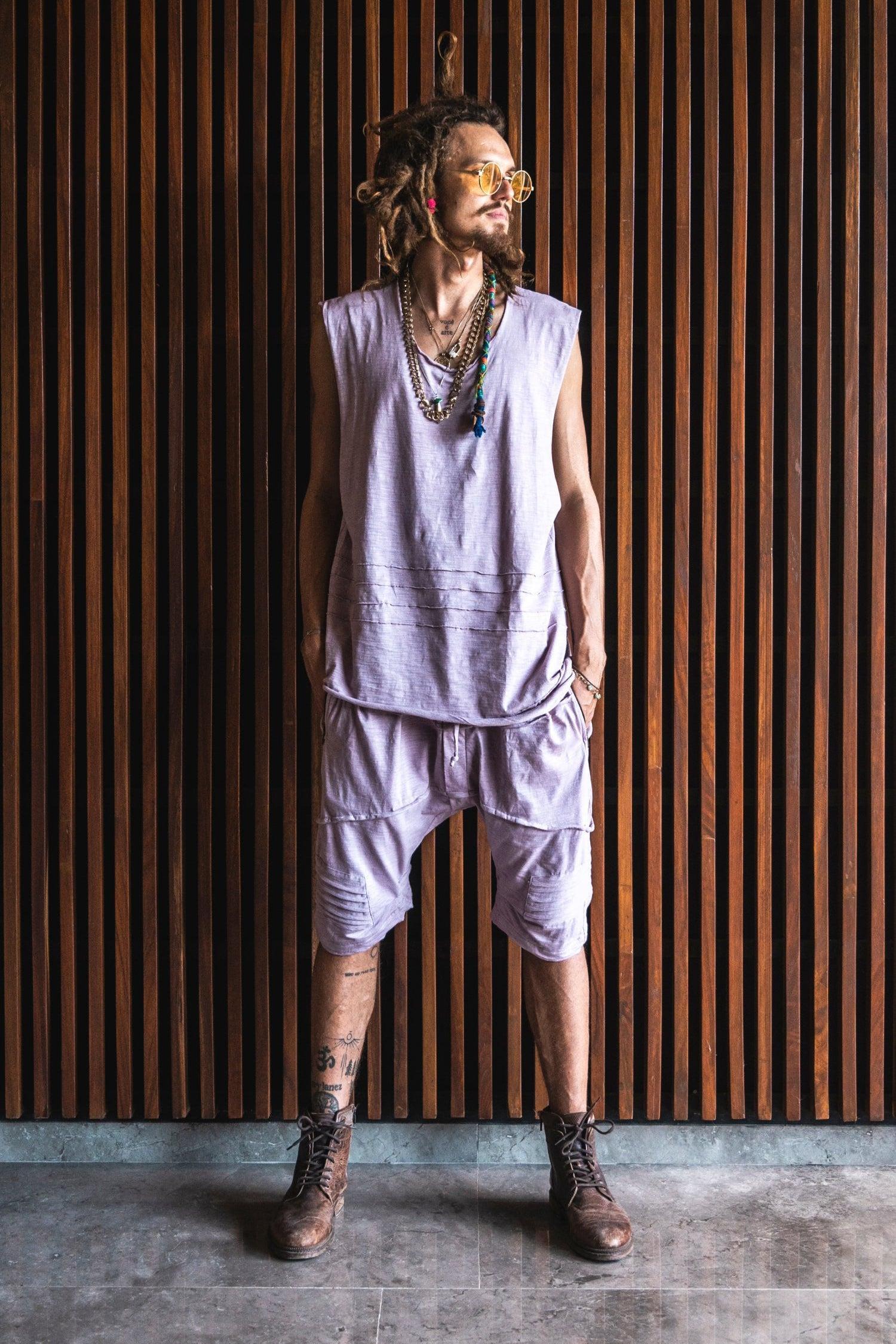 Person wearing a sleeveless top and drop crotch shorts standing against a wooden paneled wall.