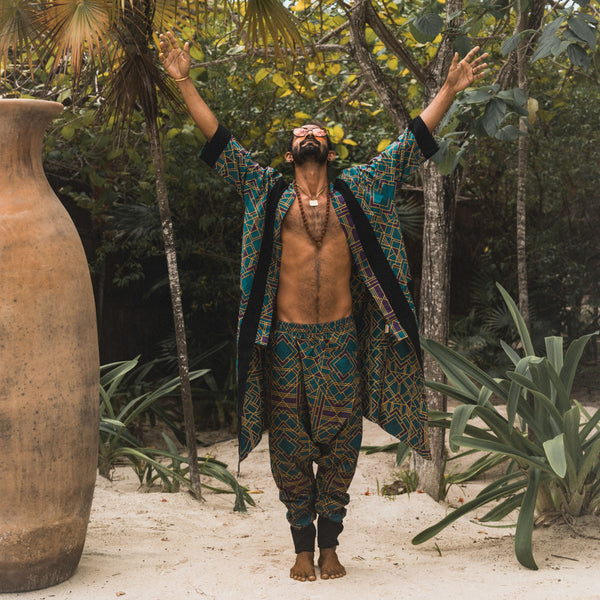 Man in patterned outfit standing in a tropical setting with plants and a large vase.