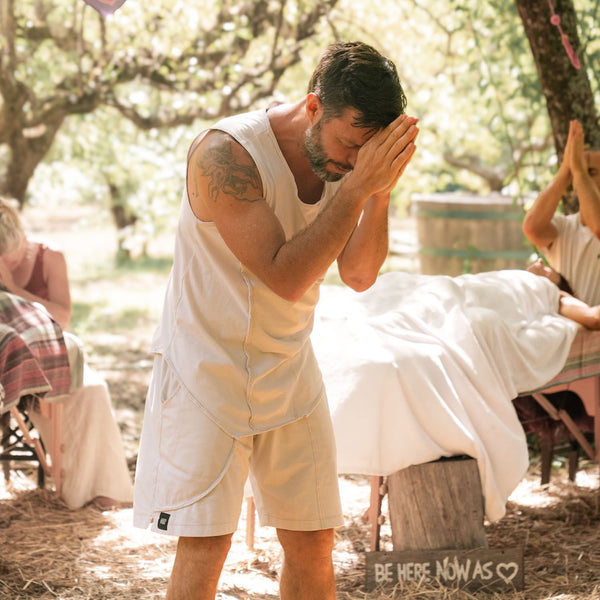 Man in a white tank top and shorts standing outdoors with trees and people in the background.