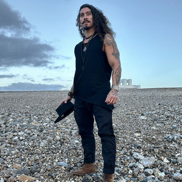 Man in black sleeveless shirt and pants standing on a rocky beach with a cloudy sky.