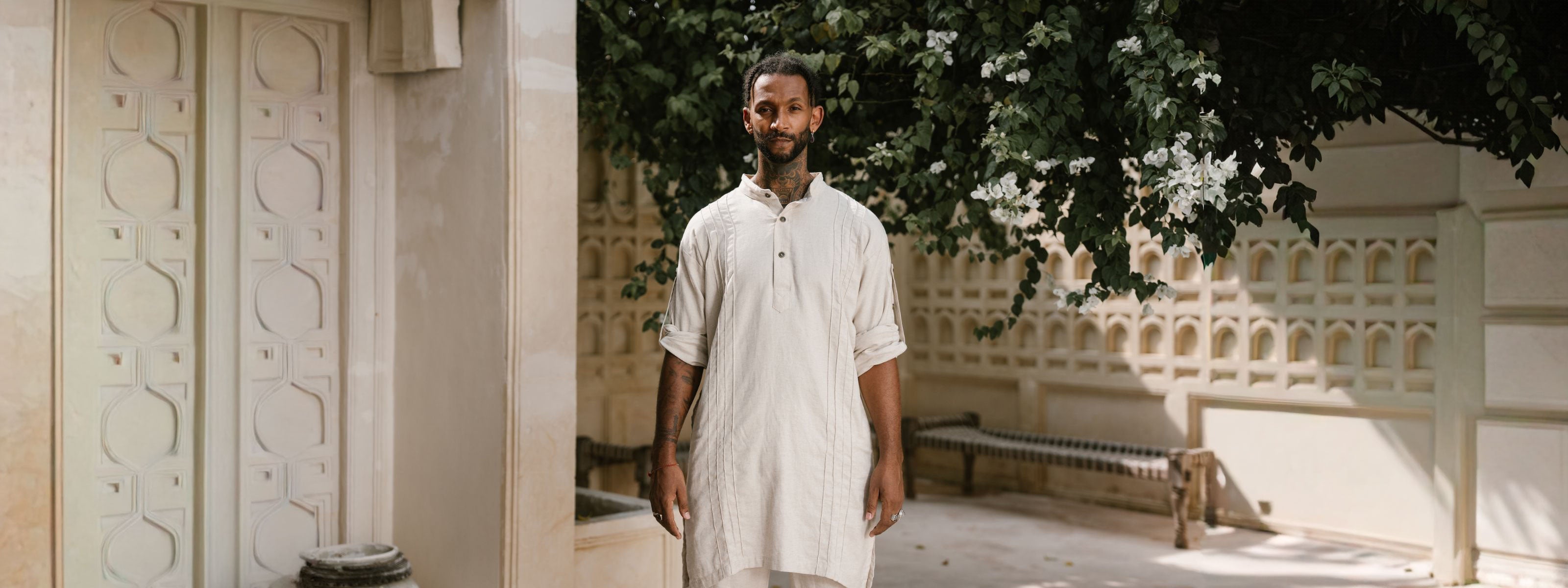 Man in a white outfit standing in a sunlit outdoor setting with architectural elements.