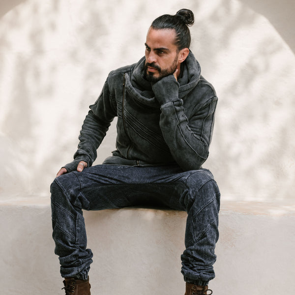 Man sitting on a white surface wearing a dark gray jacket and jeans against a neutral background