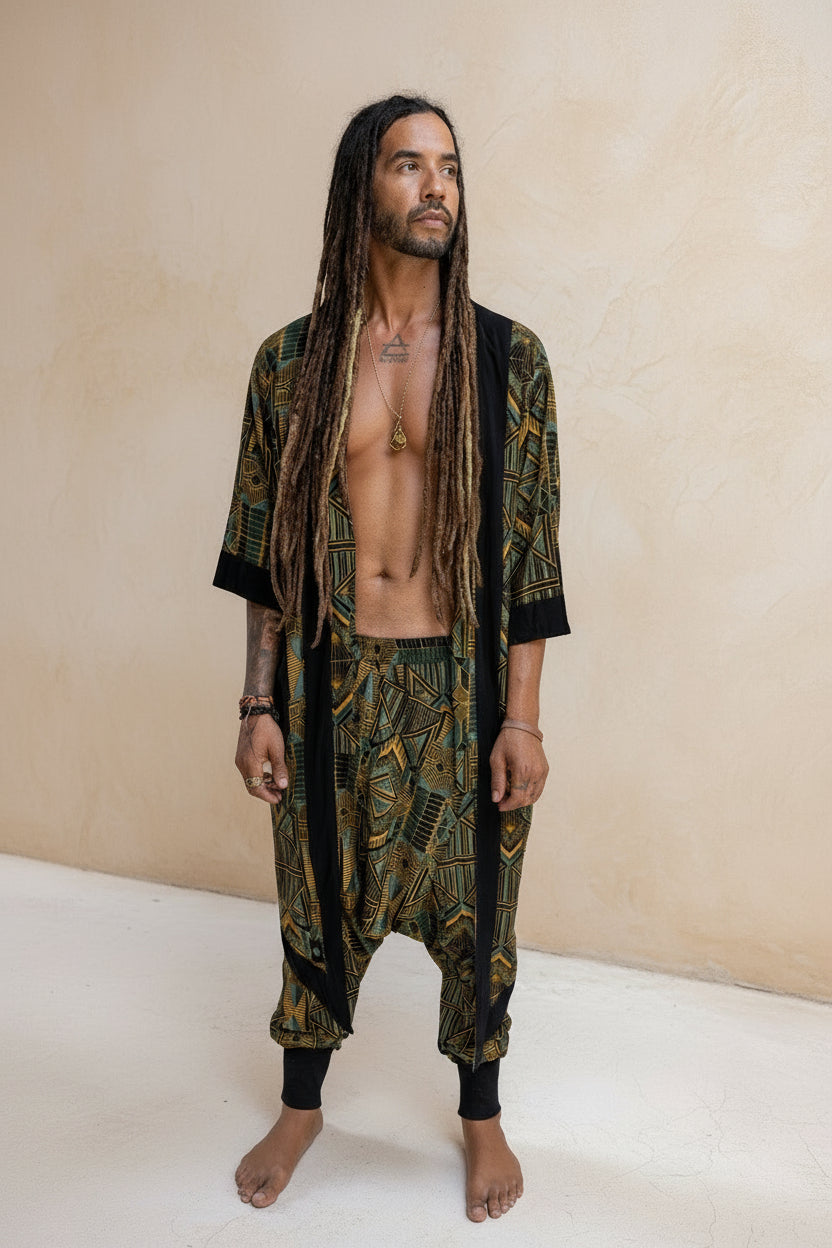 A man standing in a thatch hut wearing a blue-green geometric patterned long kimono cardigan and matching pants.